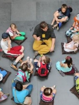 Grupa dzieci oraz osoba dorosła - przewodnik - siedzą na poduszkach rozłożonych na szarej kamiennej posadzce. Zdjęcie wykonane z perspektywy lotu ptaka.