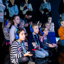 Dzieci siedzące na podłodze w słuchawkach. Za nimi rodzice/opiekunowie. Wszyscy znajdują sie w sali multimedialnej. Na podłodze wywietlają się animacje.