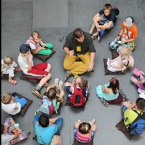 Grupa dzieci oraz osoba dorosła - przewodnik - siedzą na poduszkach rozłożonych na szarej kamiennej posadzce. Zdjęcie wykonane z perspektywy lotu ptaka.