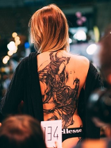 Kobieta z wytatułowanymi plecami stoi tyłem do jury.