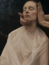 zdjęcie - siedząca kobieta w białej folii. Ma zamknięte oczy, a jej głowę trzymają dwie dłonie postaci stojącej obok.