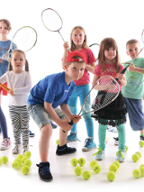 Dzieci z rakietami tenisowymi otoczone na podłodze piłkami do niego.