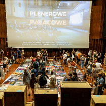 Drewniania sala z rozłożonymi plakatami na ziemi, a wśród nich ludzie wybierający je.