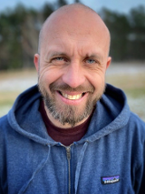 Zdjęcie Roberta Maciąga. Stoi uśmiechnięty. Głowa łysa, niebieska bluza z opuszczonym na ramiona kapturem.