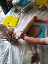 Na zdjęciu dziecięce ręce rozwałkowują glinę wałkiem na stole. Obok stoi puszka z patyczkami.