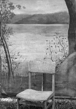 Czarno-biała fotografia przedstawia krzesło stojące na ziemi porośniętej trawą