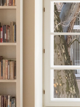 Na fotografii wnętrze Filii Sztuki Biblioteki Raczyńskich. Widać białe regały książkowe i duże okno, za którym widać fragment pnia i gałęzi platana.