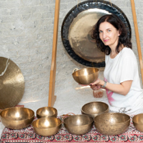 Zdjęcie przedstawia kobietę siedzącą przed dużym gongiem oraz kilkoma misami do śpiewu. Kobieta trzyma mały, czarny, prostokątny przedmiot w swojej prawej ręce. Ma na sobie białą koszulkę bez rękawów i naszyjnik. Gong jest zawieszony na suficie za kobietą. Misy do śpiewu są ułożone w półkolu przed nią. Tło stanowi biała ściana. Kobieta ma ciemne włosy i patrzy w obiektyw aparatu. Kobieta lekko się uśmiecha.