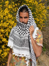 Grafika na tle miętowego koloru. Na środku znajduje się zdjęcie mężczyzny z brodą i długimi ciemnymi włosami, ubranego w kefiję.