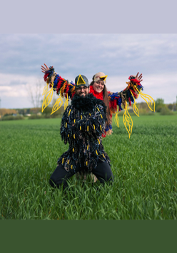 Kobieta i mężczyzna przebrani za kolorowe ptaki pozują do zdęcia. Stoją na zielonej polanie.