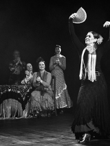 Czarno-białe zdjęcie tancerki flamenco w tanecznej pozie z wachlarzem. Na drugim placnie kilka kobiet obserwujących występ i klaszczących w dłonie.