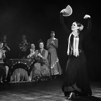 Czarno-białe zdjęcie tancerki flamenco w tanecznej pozie z wachlarzem. Na drugim placnie kilka kobiet obserwujących występ i klaszczących w dłonie.