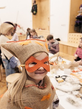 Uśmiechnięta dziewczynka w przebraniu z maską na twarzy. W tle dzieci siedzące przy stole przy pracach plastycznych.