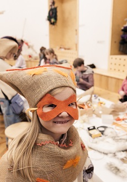 Uśmiechnięta dziewczynka w przebraniu z maską na twarzy. W tle dzieci siedzące przy stole przy pracach plastycznych.