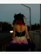 na zdjęciu widoczna postać kobiety idąca wieczorem chodnikiem ulicy, ubrana jest w sukienkę z gorsetem, z odsłoniętymi ramionami, na głowie ma woalkę z czerwonymi i czarnymi piórami i wstążkami.