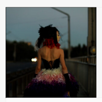 na zdjęciu widoczna postać kobiety idąca wieczorem chodnikiem ulicy, ubrana jest w sukienkę z gorsetem, z odsłoniętymi ramionami, na głowie ma woalkę z czerwonymi i czarnymi piórami i wstążkami.