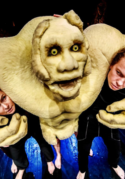 Bajkowa postać ze spektaklu, obejmująca ramionami dwóch aktorów, za nią kolejne dwie osoby.