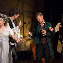 Trzech mężczyzn i jedna kobieta na scenie. Kobieta ubrana w białą suknię z wiankiem na głowie trzyma w dłoni kieliszek, mężczyźni żywo o czymś dyskutują.