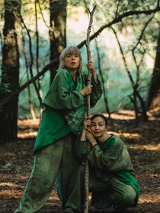 Dwie kobiety w lesie, ubrane w zielone atroje, trzymają cienki pień drzewa.