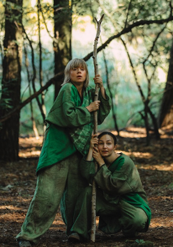 Dwie kobiety w lesie, ubrane w zielone atroje, trzymają cienki pień drzewa.