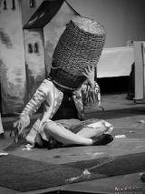 Czarno-białe zdjęcie osoby siedzącej na podłodze ze skrzyżowanymi nogami z dużym koszum na głowie; kosz całkowicie zakrywa twarz.