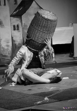 Czarno-białe zdjęcie osoby siedzącej na podłodze ze skrzyżowanymi nogami z dużym koszum na głowie; kosz całkowicie zakrywa twarz.