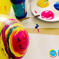 Kolorowe zdjęcie żółtego balona częściowo pomalowanego w kolorowe okręgi. Na drugim planie papierowy talerz z trzema kolorowymi plamami, o talerz oparty jest pędzelek.