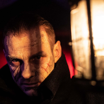 Twarz mężczyzny w półmroku, na którą częściowo pada światło. Na twarzy widoczne blizny.