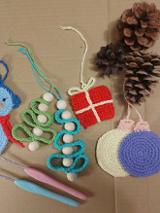 Na zdjęciu kilka prac z kolorowej włóczki wykonanych szydełkiem, takich jak niebieski bałwanek, bombki, choinki. Na zdjęciu też trzy szyszki i dwa szydełka.
