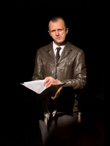 Na zdjęciu mężczyzna w brązowej skórzanej kurtce. W dłoniach trzyma teczkę, patrzy w obiektyw.