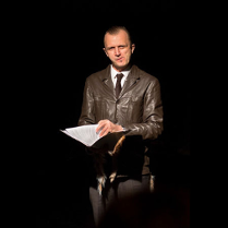 Na zdjęciu mężczyzna w brązowej skórzanej kurtce. W dłoniach trzyma teczkę, patrzy w obiektyw.