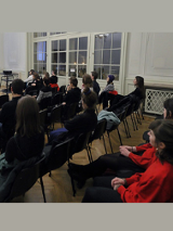 Zdjęcie przedstawia grupę osób, które siedzą na krzesłach i słuchają wykładu