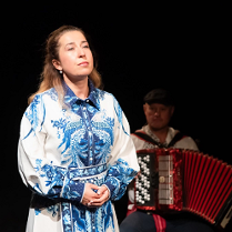 Zdjęcie kobiety w biało-niebieskiej sukience, która patrzy w dal. W tle mężczyzna grający na akordeonie.