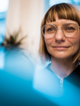 Kobieta w okularach i krótkich włosach. Zdjęcie w błękitno-beżowej kolorystyce.