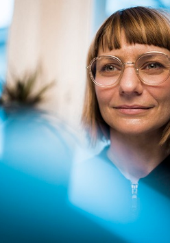 Kobieta w okularach i krótkich włosach. Zdjęcie w błękitno-beżowej kolorystyce.