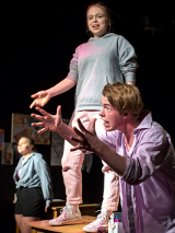 na zdjęciu troje młodych aktorów, na pierwszym planie młody mężczyzna pochylony do przodu gestykuluje rękami, za nim uśmiechnięta dziewczyna stoi na stole, w tle kolejna dziewczyna stoi oparta o stół