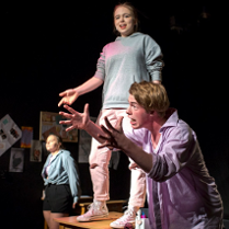 na zdjęciu troje młodych aktorów, na pierwszym planie młody mężczyzna pochylony do przodu gestykuluje rękami, za nim uśmiechnięta dziewczyna stoi na stole, w tle kolejna dziewczyna stoi oparta o stół