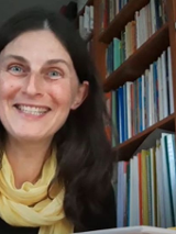 Uśmiechająca się kobieta w ciemnych włosach i zółtym szaliku. Za nią regały pełne książek.