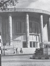 Zdjęcie czarno-białe. Ulica, a na niej autobus i pieszy stojacy pod drzewem. W tle okrągły budynek.