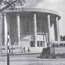 Zdjęcie czarno-białe. Ulica, a na niej autobus i pieszy stojacy pod drzewem. W tle okrągły budynek.