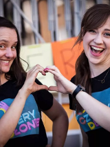 Zdjęcie przedsawie dwie uśmiechnięte kobiety, które razem, przy pomoy dłoni, tworzą kształt serduszka.