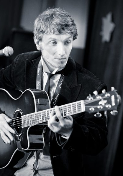 zdjecie wykonawcy - mężczyzna stojący na scenie przy mikrofonie, grający na gitarze
