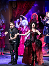 Zdjęcie ze spektaklu - na scenie aktorzy w różnorodnych strojach; na pierwszym planie aktorka w długiej balowej sukni, obok niej mężczyzna w skórzanych spodniach, skórzanej kamizelce i T-shircie.