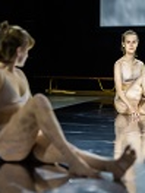 Zdjęcie dwóch młodych kobiet ubranych w biała bieliznę, siedzących na podłodze, każda z podkurczoną nogą. W tle drewniana ławka gimnastyczna.