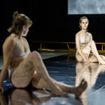 Zdjęcie dwóch młodych kobiet ubranych w biała bieliznę, siedzących na podłodze, każda z podkurczoną nogą. W tle drewniana ławka gimnastyczna.