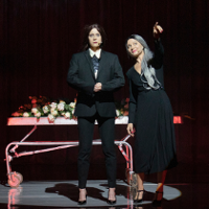 Na pierwszym planie dwie stojące postaci ubrane na czarno, za nimi wysokie metalowe łóżko na kółkach okryte kwiatami