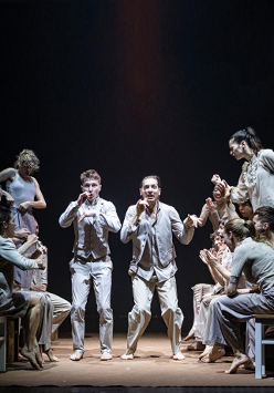 Grupa tancerzy w jasnych ubraniach podczas spketaklu: w środku dwóch mężczyzn stojących na ugiętych nogach, po bokach po kilka osób stojących bokiem lub siedzących na taboretach.