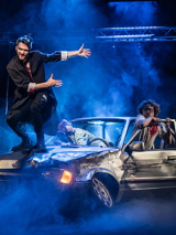 Zdjęcie przedstawiające trzy osoby wysiadające ze zniszczonego samochodu oraz jednego mężczyznę stojącego na masce samochodu.