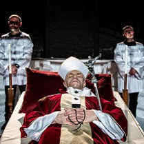 Mężczyzna ubrany w szaty papieskie leży na katafalku, pod głową ma purpurową poduszkę; za nim stoją dwaj mężczyźni w komżach.