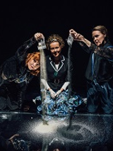 Trzy kobiety, dwie z nich wysypują piasek z woreczków na czarną powierzchnię. Czarne tło.